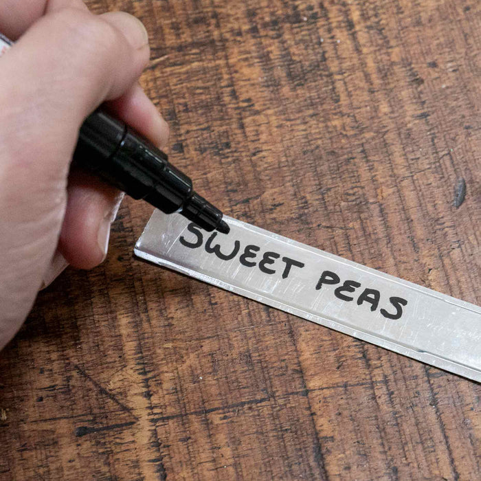 Hand writing 'SWEET PEAS' on a metal tag with a marker on a wooden surface.