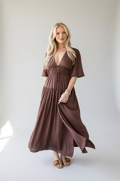 A blonde woman modeling a brown, knee-length maxi dress with short flutter sleeves, a deep V-neckline, and a gathered waist, standing against a neutral light gray studio background.