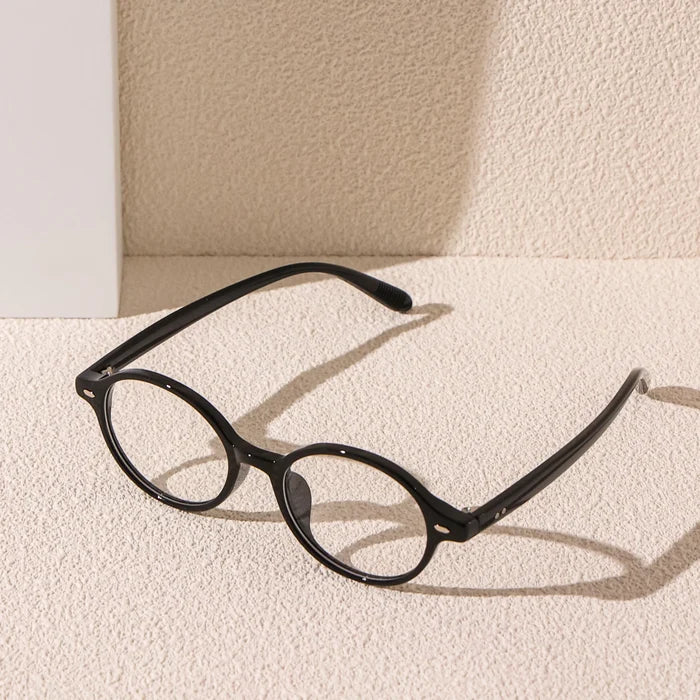 A pair of EyeCandys Chase Round Oval Glasses with clear lenses rests on a textured beige surface, casting soft shadows against a light-colored wall in the background.