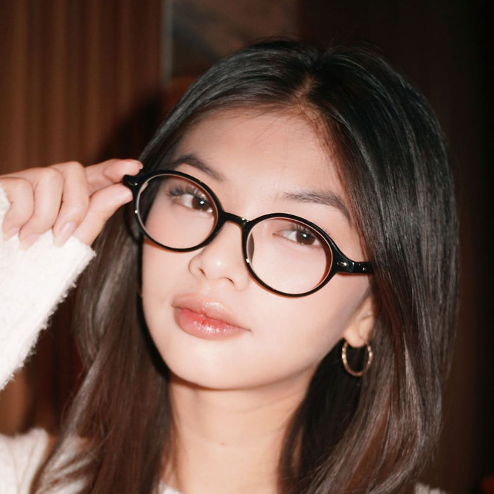 A young woman with long, straight brown hair wears EyeCandys Chase Round Oval glasses and gold hoop earrings. She slightly smiles, holding her glasses with one hand and looking directly at the camera.
