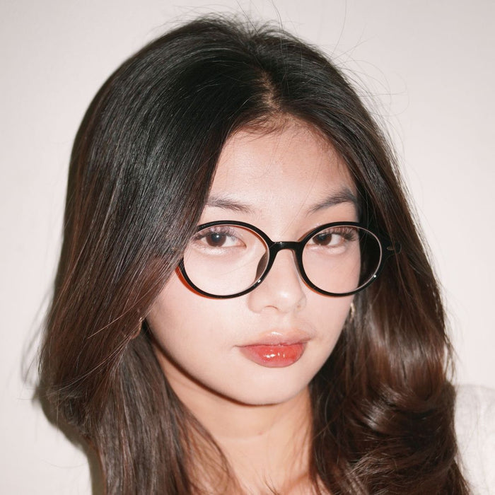 A person with long dark hair wears EyeCandys Chase Round Oval eyeglasses, looking at the camera with a neutral expression against a plain light background.