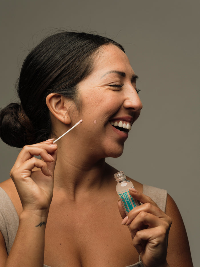 Woman applying acne treatment to her face with a cotton swab against a neutral background