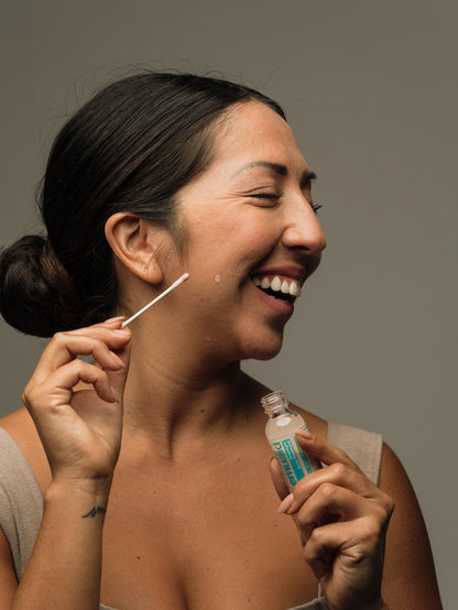 Woman applying acne treatment to her face with a cotton swab against a neutral background