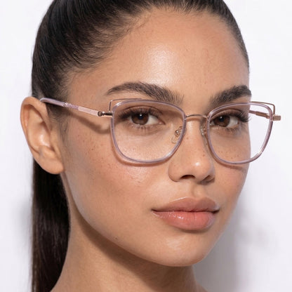 A woman with long dark hair in a ponytail wears EyeCandys Kaylee Cat Eye oversized light pink glasses, showing smooth skin, natural makeup, and freckles. She has a neutral expression and stands against a plain white background.