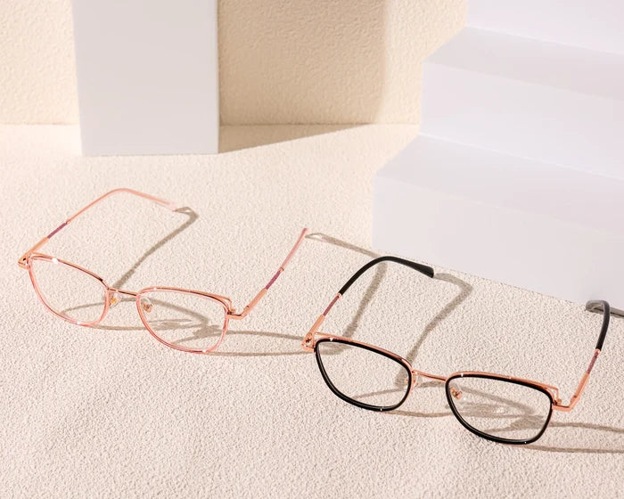Two pairs of EyeCandys Kaylee Cat Eye glasses with thin metal frames—one in rose gold, the other in rose gold and black—rest on a textured beige surface, highlighted by white geometric blocks for a chic, modern look.