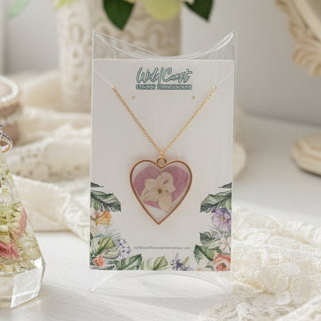 Gold heart pendant with pressed white flower in resin on a floral card, a preserved-flower keepsake jewelry set.