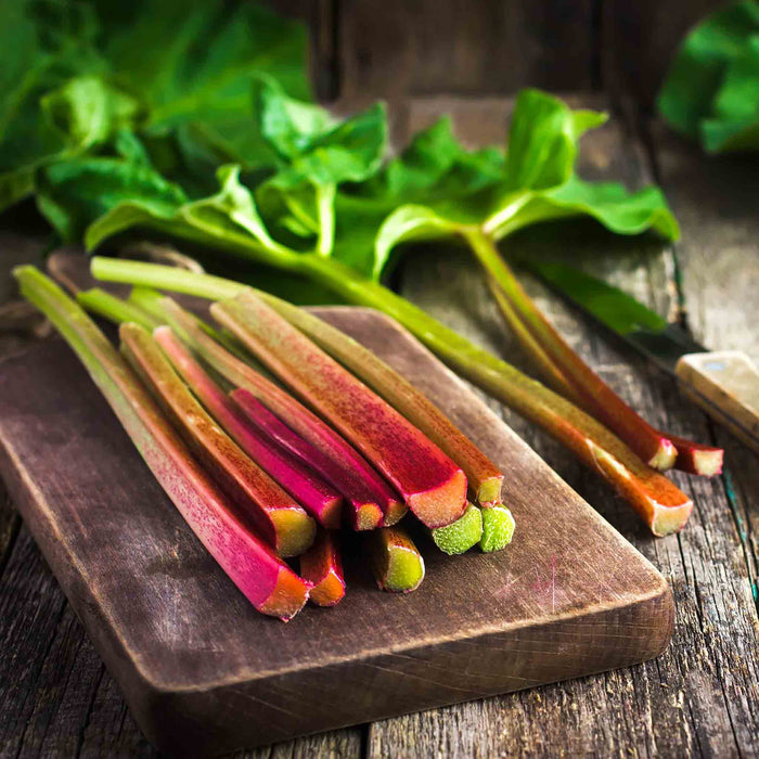 Fresh rhubarb stalks on a wooden cutting board with leaves in the background, indicating the product is rhubarb plants suitable for culinary use.