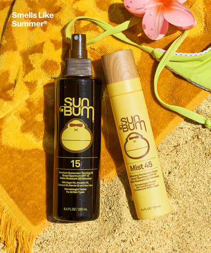 Sun Bum SPF 15 Tanning Oil + Mist 45 bottles on yellow towel beside sandy beach and green bikini top