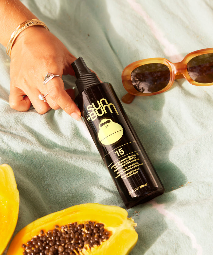 Hand holding Sun Bum SPF 15 Tanning Oil + Mist 45 bottle on a towel with sunglasses and papaya fruit nearby