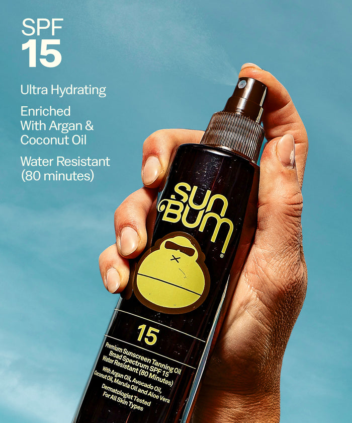 Hand holding Sun Bum SPF 15 Tanning Oil + Mist 45 bottle with hydrating argan and coconut oils visible