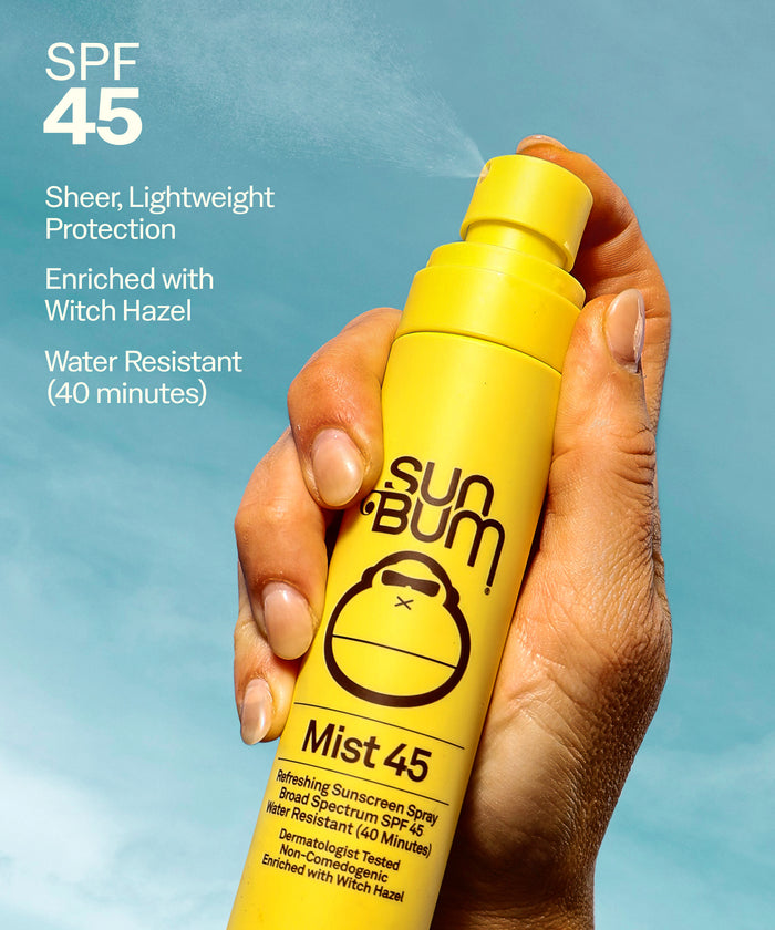Hand holding Sun Bum SPF 15 Tanning Oil + Mist 45 yellow spray bottle misting sunscreen against sky background