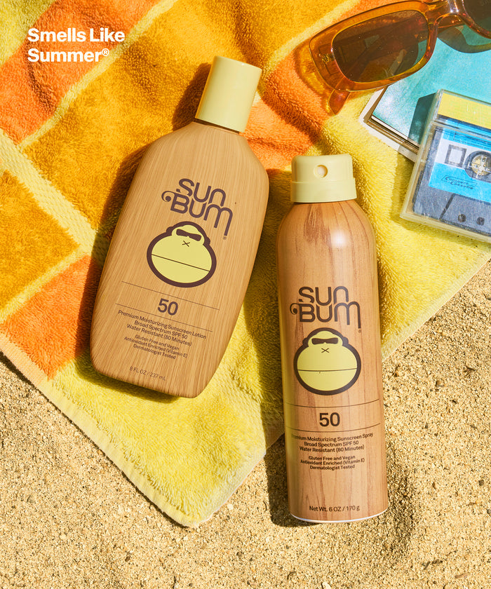 Sun Bum SPF 50 Spray + Lotion bottles on a colorful beach towel with sunglasses and a cassette tape nearby