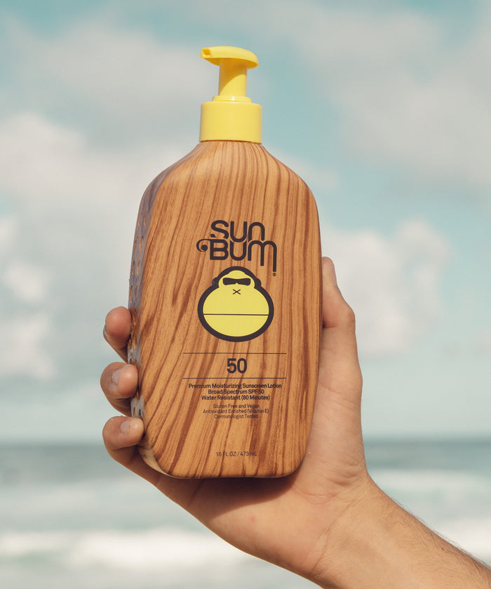 Hand holding Sun Bum XL SPF 50 Lotion + Face and Body stick 40 with wooden texture and yellow pump against sky background