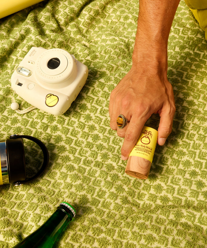 Hand holding Sun Bum XL SPF 50 Lotion + Face and Body stick 40 on green patterned surface near camera and water bottle